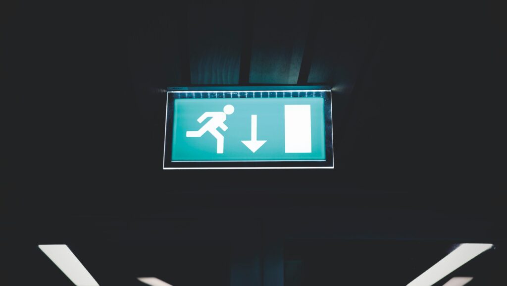 Brightly lit emergency exit sign with arrow indicating direction, set against a dark backdrop.